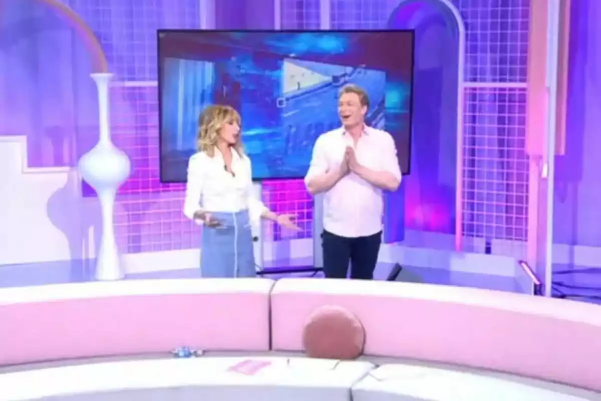 Emma García y Richard Pena de pie en Fiesta con decoración en tonos rosados y morados, una pantalla grande detrás de ellos y un sofá curvo en primer plano. Emma García y Richard Pena de pie en Fiesta con decoración en tonos rosados y morados, una pantalla grande detrás de ellos y un sofá curvo en primer plano.