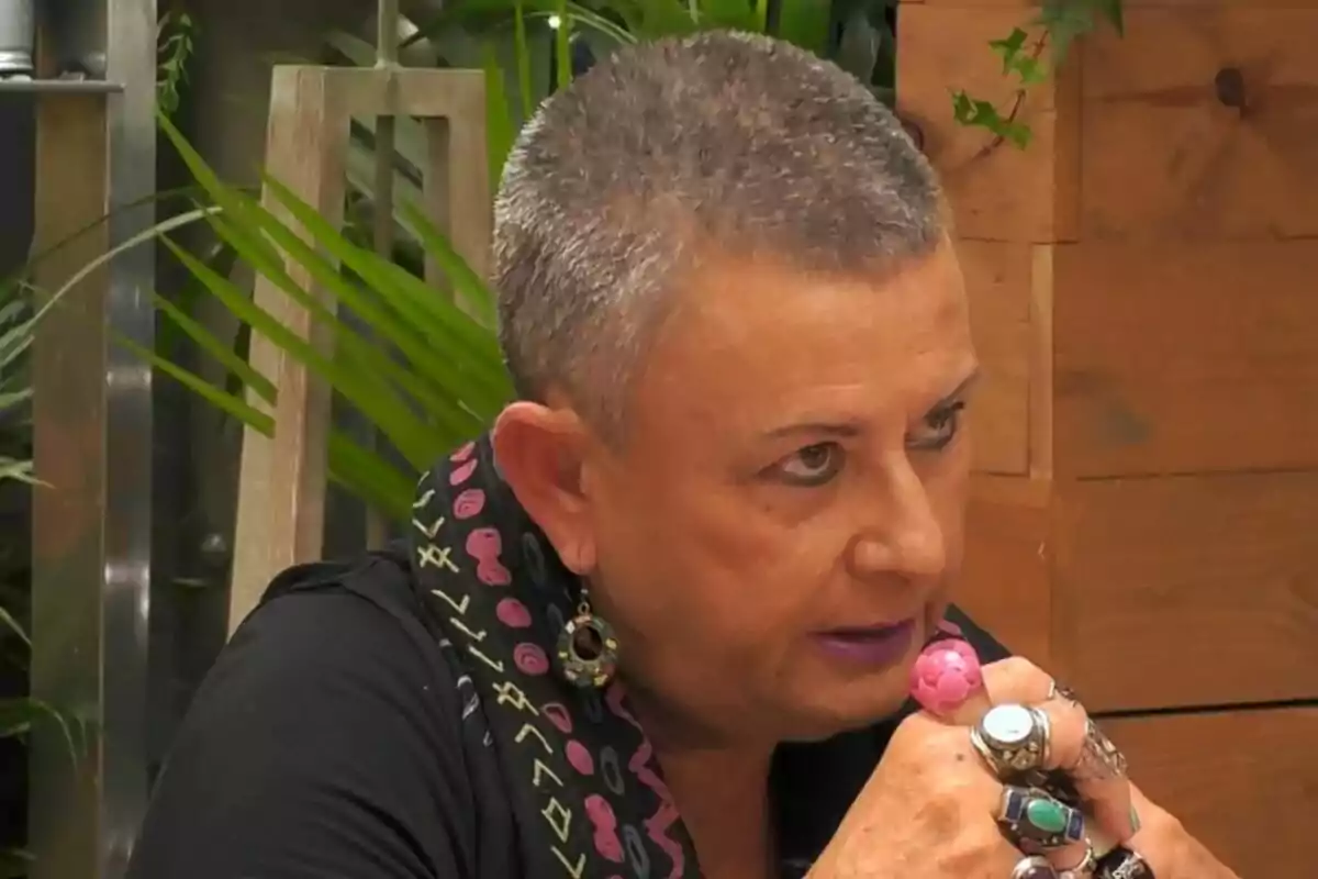 Una persona con cabello corto y canoso, usando un pañuelo con estampado colorido, pendientes grandes y varios anillos, está sentada en un entorno con plantas y madera en First Dates. Una persona con cabello corto y canoso, usando un pañuelo con estampado colorido, pendientes grandes y varios anillos, está sentada en un entorno con plantas y madera en First Dates.