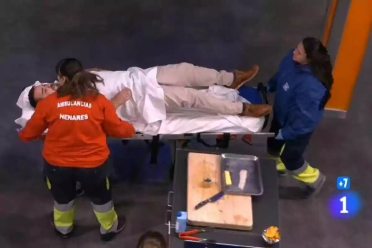 Flores (concursante de MasterChef) está acostado en una camilla siendo atendido por personal de emergencias mientras hay una mesa con utensilios de cocina cerca. Flores (concursante de MasterChef) está acostado en una camilla siendo atendido por personal de emergencias mientras hay una mesa con utensilios de cocina cerca.