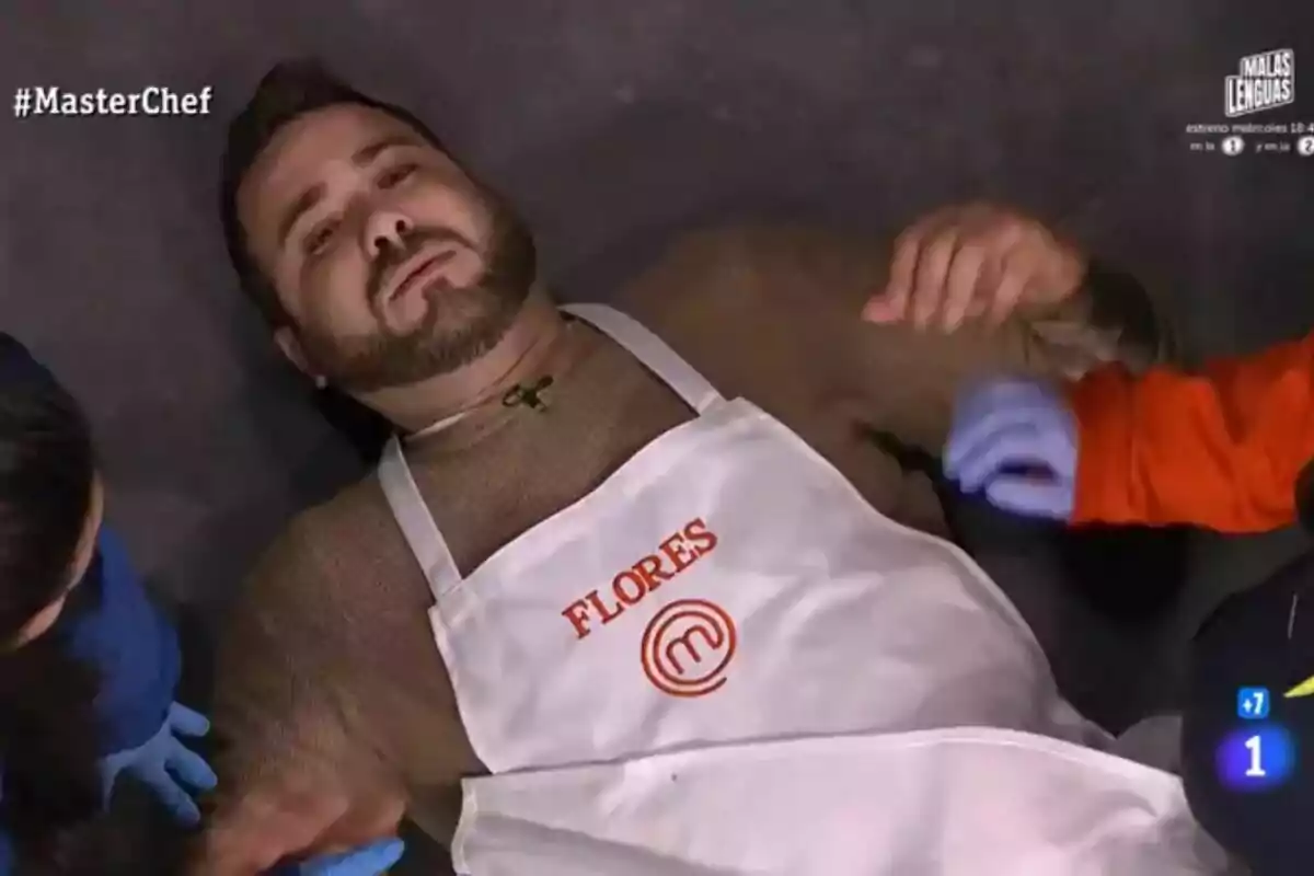 Un participante de MasterChef está acostado en el suelo con un delantal que tiene el nombre "Flores", mientras otras personas con guantes lo asisten.