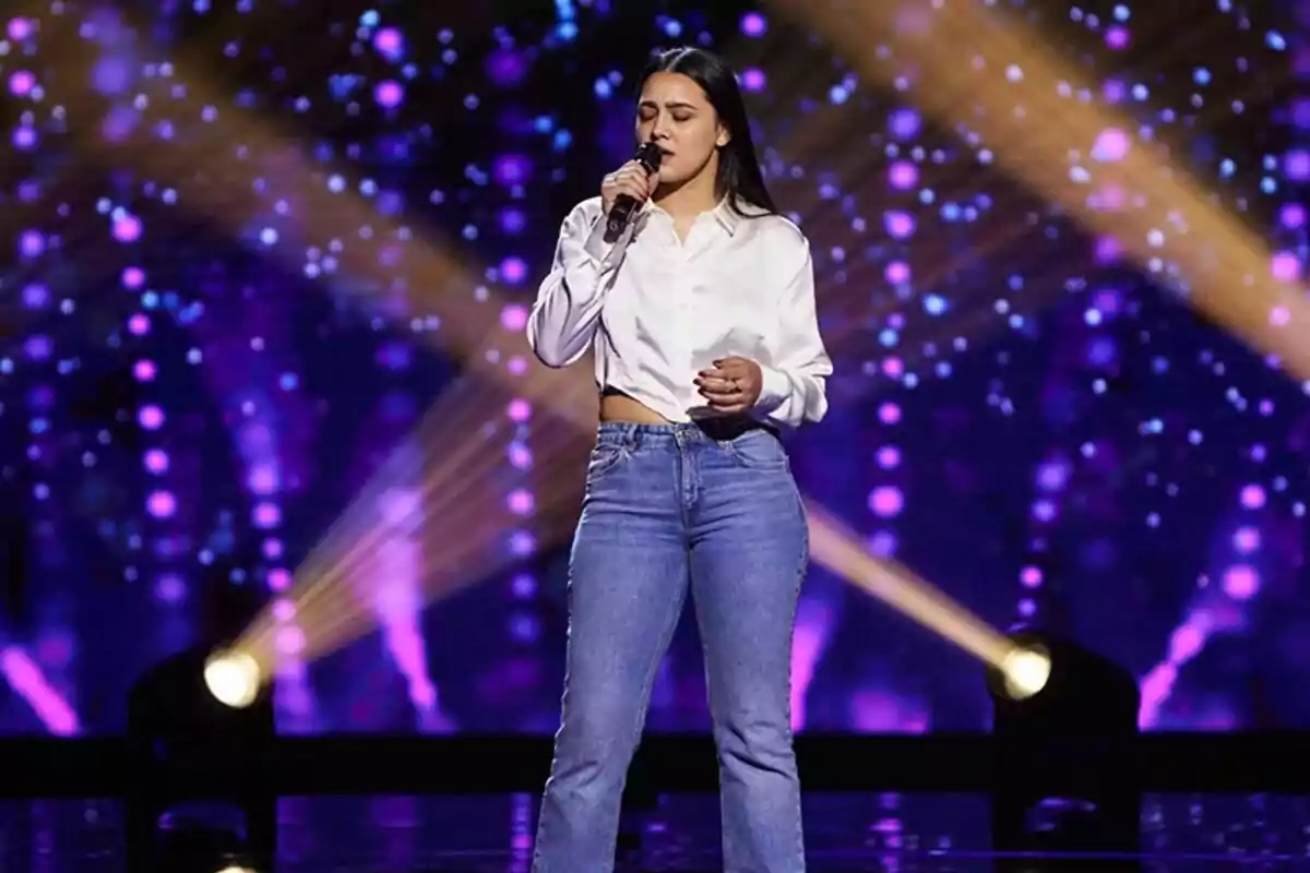 Una mujer canta en un escenario de La Voz iluminado con luces moradas y doradas. Una mujer canta en un escenario de La Voz iluminado con luces moradas y doradas.