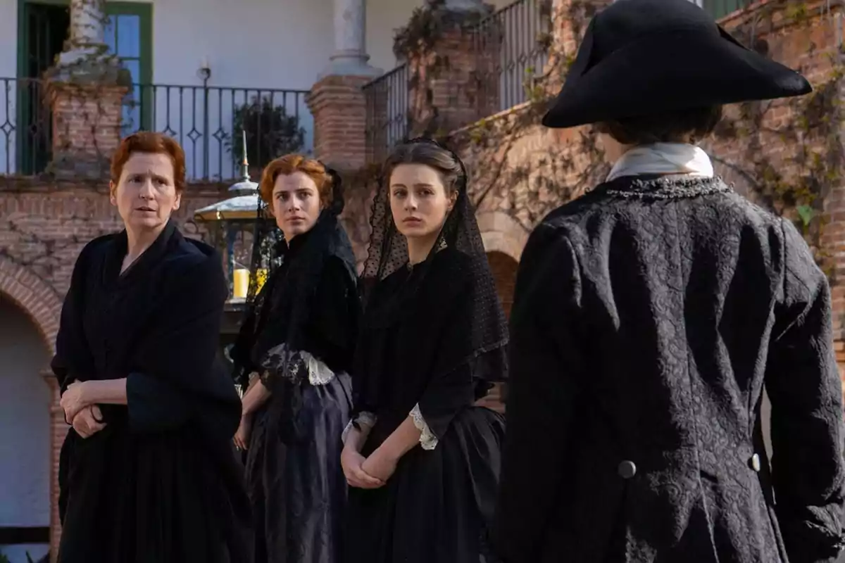 Escena de Valle Salvaje, amb Bàrbara, Adriana i Isabel, vestides de negre miren una persona amb barret en un pati amb parets de maó i una barana de ferro. Escena de Valle Salvaje, amb Bàrbara, Adriana i Isabel, vestides de negre miren una persona amb barret en un pati amb parets de maó i una barana de ferro.