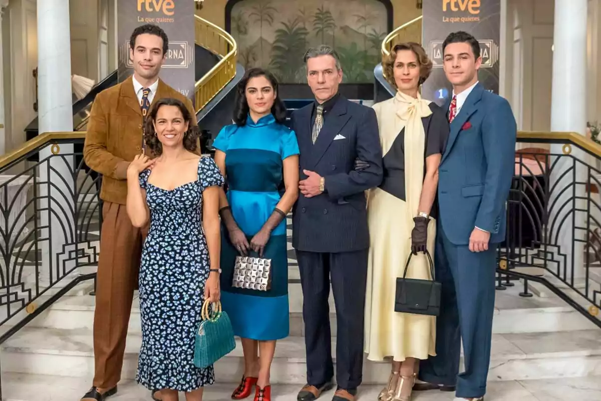 Un grupo de personas elegantemente vestidas posando en una escalera con una decoración de fondo que incluye barandales dorados y un mural en La Moderna Un grupo de personas elegantemente vestidas posando en una escalera con una decoración de fondo que incluye barandales dorados y un mural en La Moderna