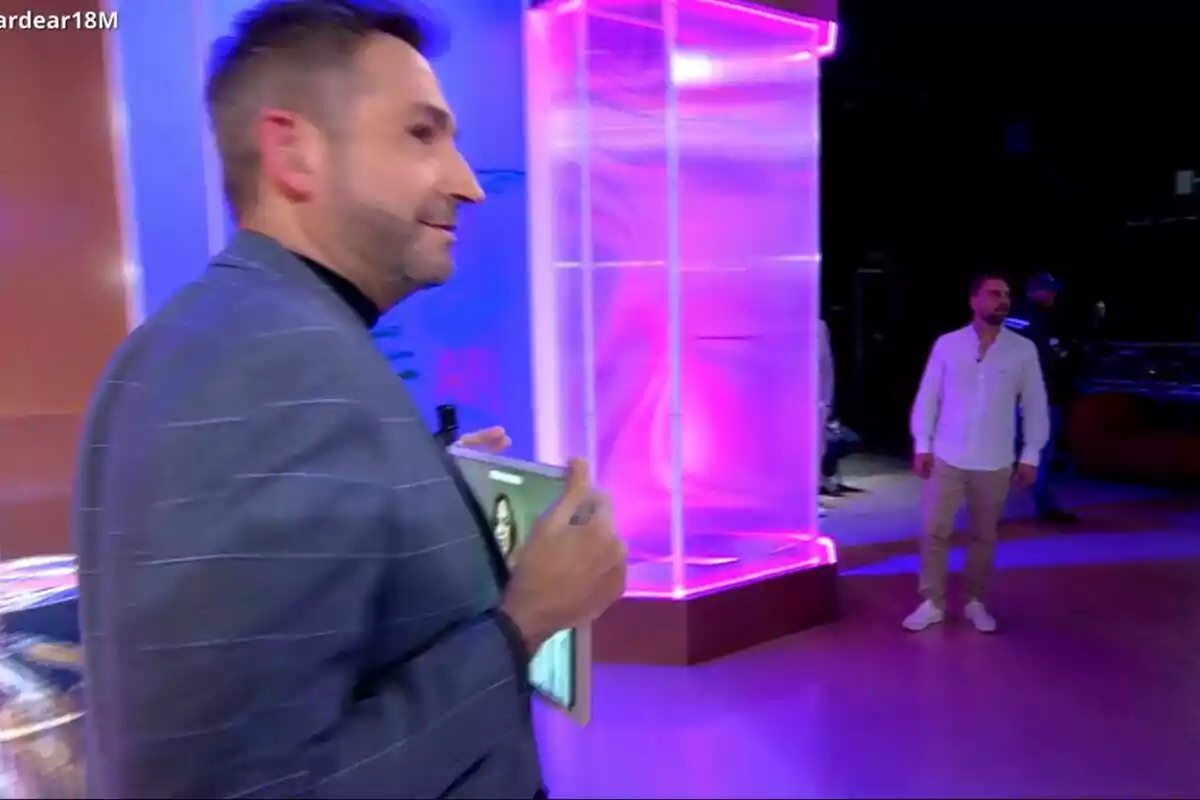 Frank Blanco con chaqueta sostiene una tableta mientras camina en un estudio de Tardear iluminado con luces de colores, al fondo se ve a José María Almoguera de pie. Frank Blanco con chaqueta sostiene una tableta mientras camina en un estudio de Tardear iluminado con luces de colores, al fondo se ve a José María Almoguera de pie.