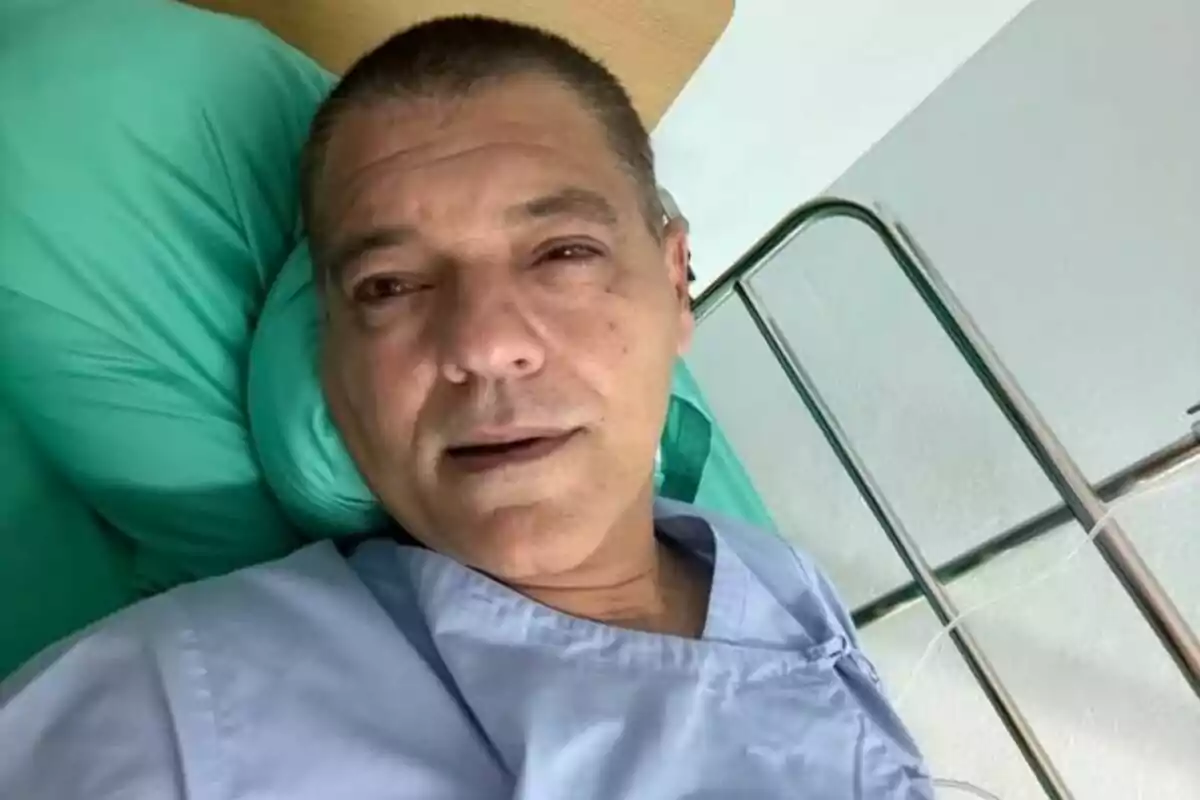 Frank Cuesta recostado en una cama de hospital con una bata azul y una almohada verde.