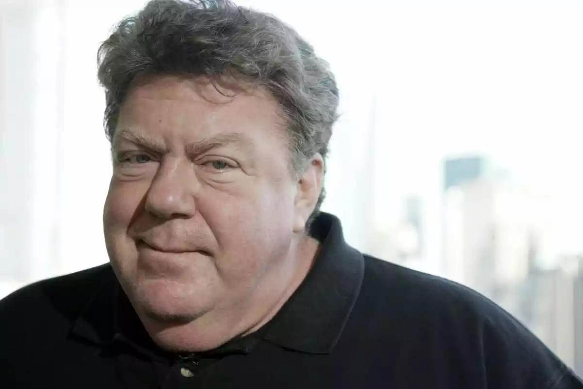 George Wendt amb cabell arrissat i una expressió neutral porta una camisa fosca amb un fons desenfocat.