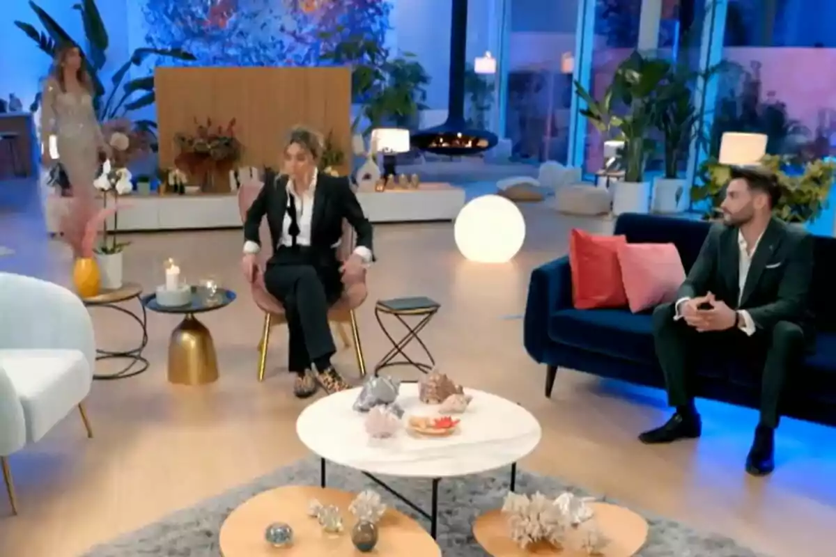 Un elegante salón moderno con decoración contemporánea en La Isla de las Tentaciones, donde tres personas están presentes: Alba de pie al fondo, Sandra Barneda en una silla y Gerard en un sofá, rodeados de plantas y una iluminación cálida. Un elegante salón moderno con decoración contemporánea en La Isla de las Tentaciones, donde tres personas están presentes: Alba de pie al fondo, Sandra Barneda en una silla y Gerard en un sofá, rodeados de plantas y una iluminación cálida.
