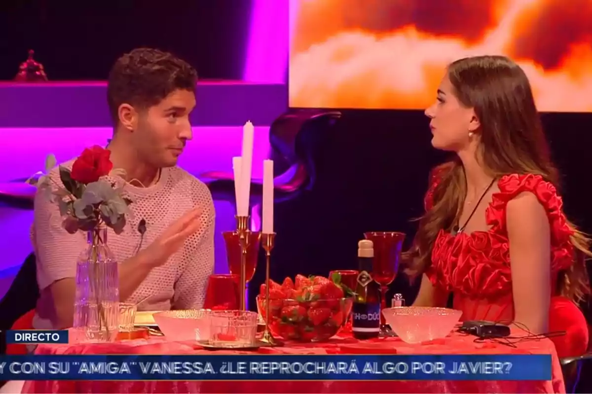 Álex Ghita y Maica conversan en un set de televisión decorado con tonos rojos y morados, con una mesa adornada con velas, una rosa y fresas. Captura de 'GH Dúo'
