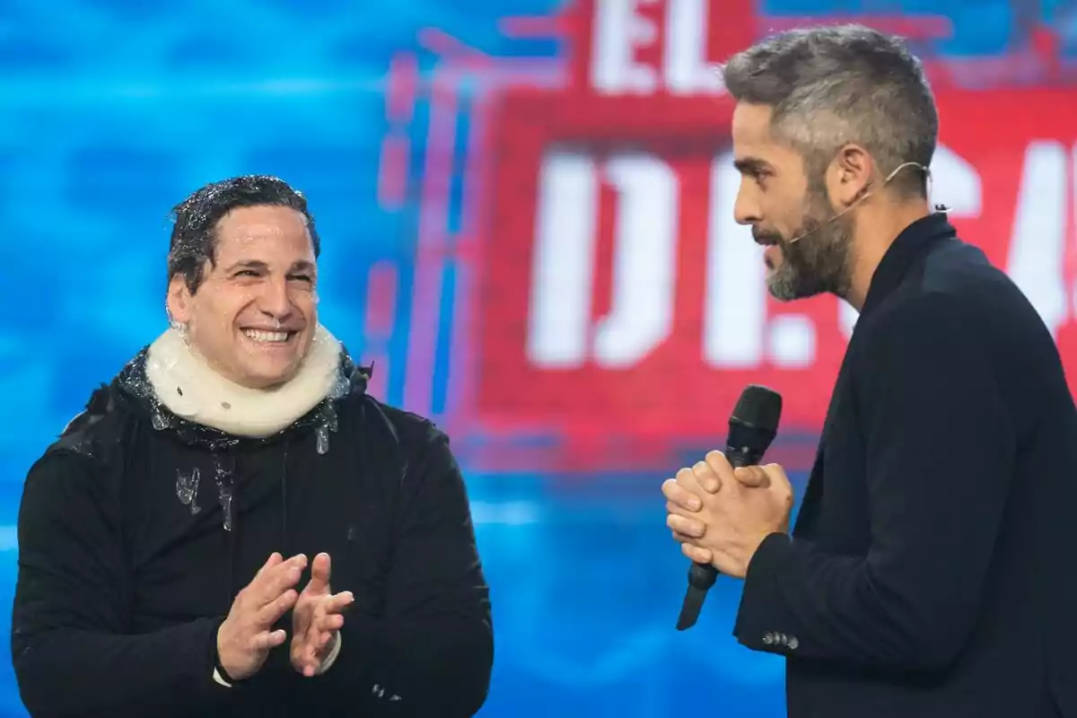 Gotzon Mantuliz y Roberto Leal en un escenario de televisión una de ellas con un micrófono y la otra con un collarín y hielo en la cabeza sonríe mientras aplaude en El Desafío. Gotzon Mantuliz y Roberto Leal en un escenario de televisión una de ellas con un micrófono y la otra con un collarín y hielo en la cabeza sonríe mientras aplaude en El Desafío.