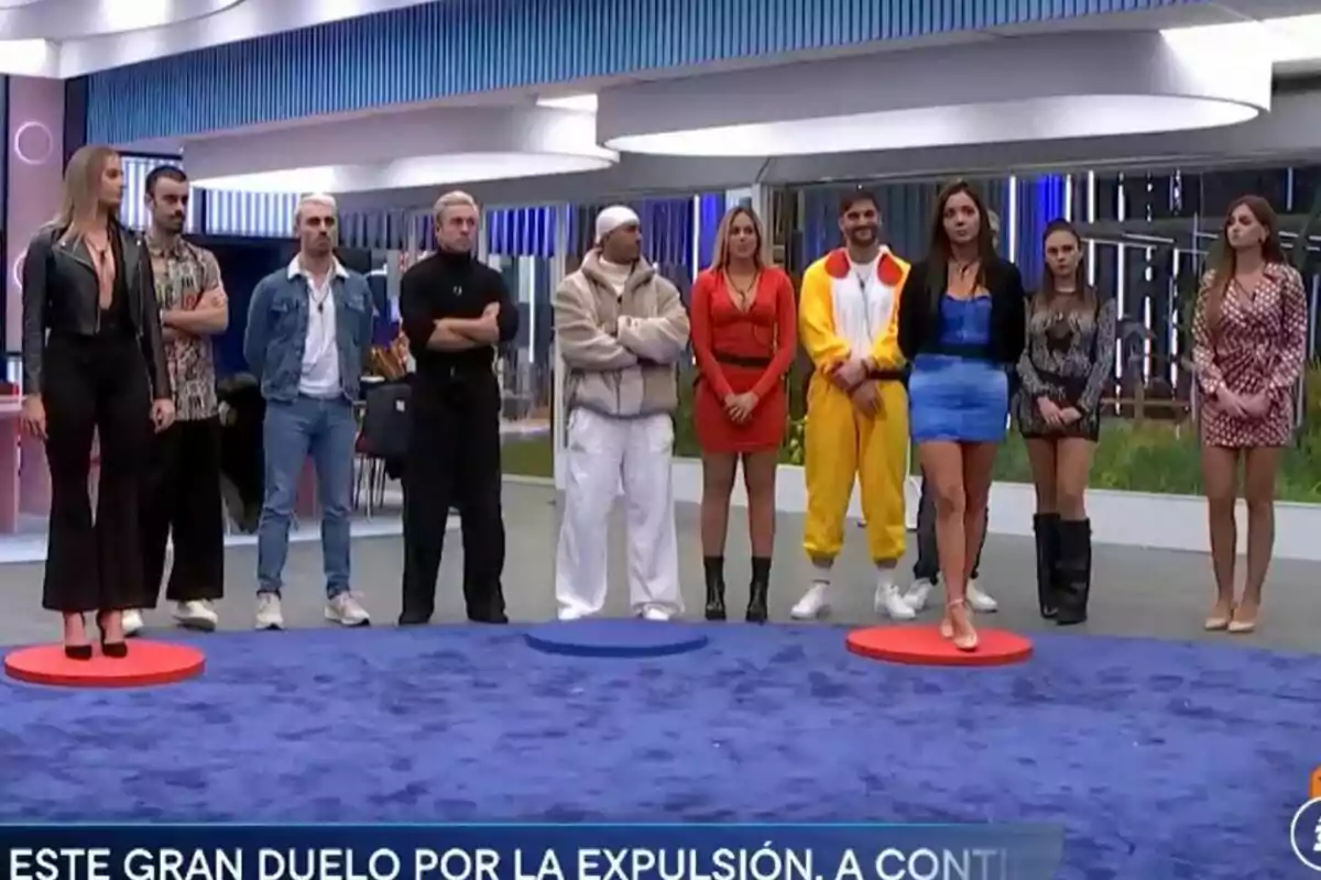 Un grupo de concursantes de Gran Hermano en pie en la casa, algunas sobre círculos de colores en el suelo, con un letrero en la parte inferior que menciona un duelo por la expulsión. Un grupo de concursantes de Gran Hermano en pie en la casa, algunas sobre círculos de colores en el suelo, con un letrero en la parte inferior que menciona un duelo por la expulsión.