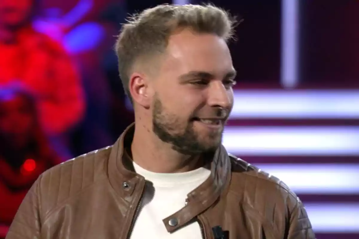 José María Almoguera con chaqueta de cuero marrón y camiseta blanca sonríe mientras mira hacia un lado en un entorno con luces de fondo. Captura de 'Gran Hermano' José María Almoguera con chaqueta de cuero marrón y camiseta blanca sonríe mientras mira hacia un lado en un entorno con luces de fondo. Captura de 'Gran Hermano'
