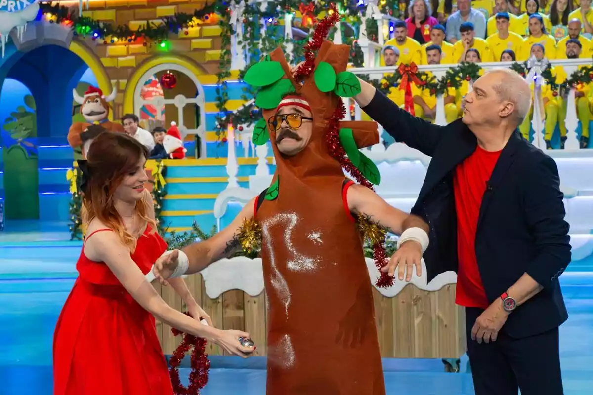 Wilbur disfrazado de árbol está en El Grand Prix decorado con motivos navideños, acompañado por Cristinini de vestido rojo y Ramón García de traje oscuro, mientras un público vestido de amarillo observa desde las gradas. Wilbur disfrazado de árbol está en El Grand Prix decorado con motivos navideños, acompañado por Cristinini de vestido rojo y Ramón García de traje oscuro, mientras un público vestido de amarillo observa desde las gradas.