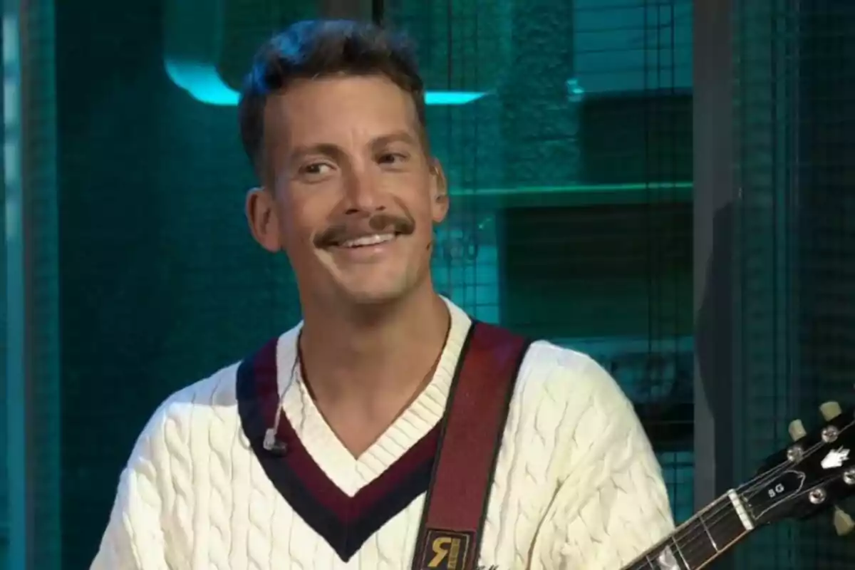 Grison somrient amb bigoti i suèter blanc sosté una guitarra a La Revuelta. Grison somrient amb bigoti i suèter blanc sosté una guitarra a La Revuelta.