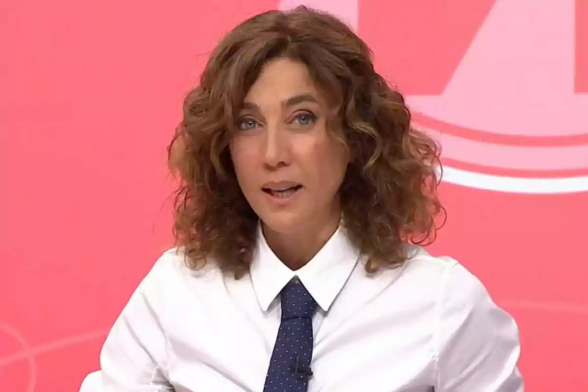 Dona amb cabell arrissat i camisa blanca amb corbata blava sobre fons rosa.