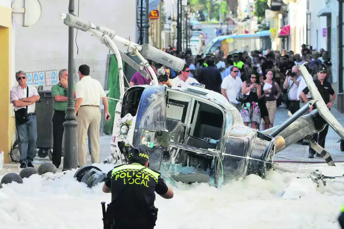 Un helicòpter accidentat jeu en un carrer envoltat d'escuma mentre un policia local observa l'escena i diverses persones s'agrupen al voltant.