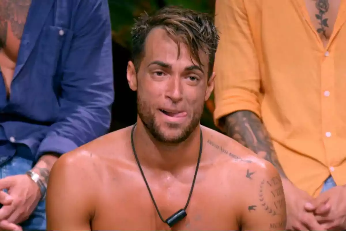 Montoya sin camisa con tatuajes visibles en los hombros y pecho, rodeado de otras personas con camisas de colores en La Isla de las Tentaciones. Montoya sin camisa con tatuajes visibles en los hombros y pecho, rodeado de otras personas con camisas de colores en La Isla de las Tentaciones.