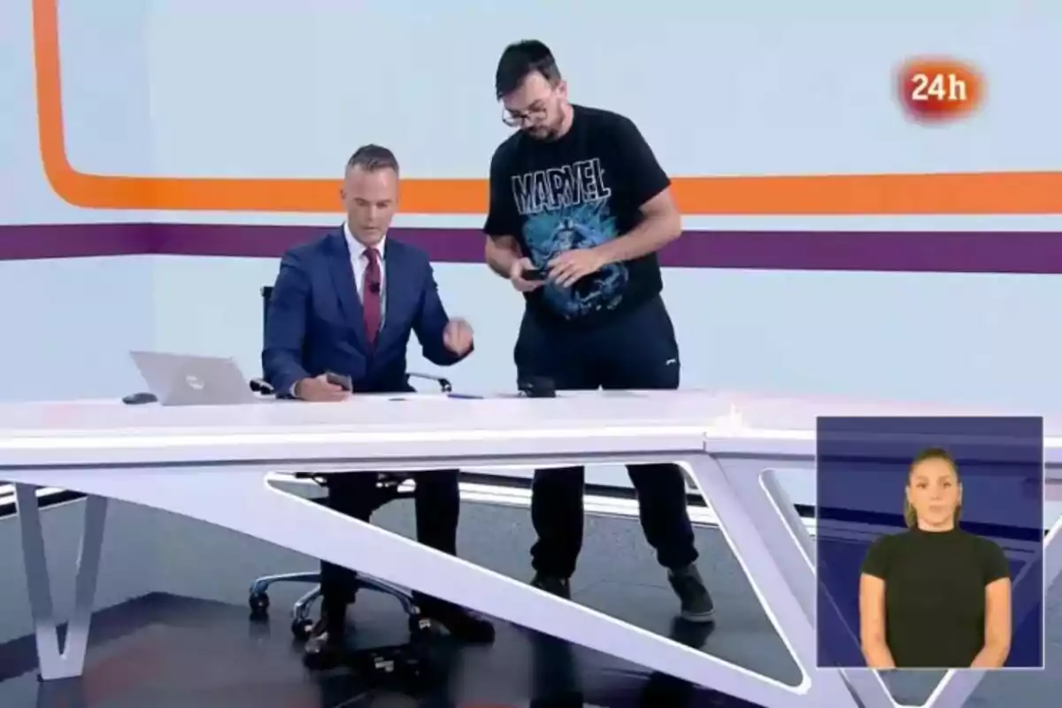 Igor Gómez en el Telediario está sentado en un escritorio con un portátil mientras un técnico de pie a su lado sostiene un dispositivo móvil y en la esquina inferior derecha hay una intérprete de lenguaje de señas. Igor Gómez en el Telediario está sentado en un escritorio con un portátil mientras un técnico de pie a su lado sostiene un dispositivo móvil y en la esquina inferior derecha hay una intérprete de lenguaje de señas.