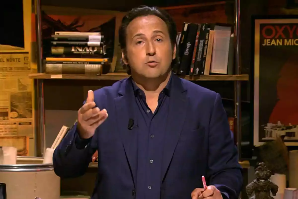 Captura de Iker Jiménez con traje azul oscuro está hablando y gesticulando con las manos, mientras está sentado frente a una estantería llena de libros en el final de Cuarto Milenio