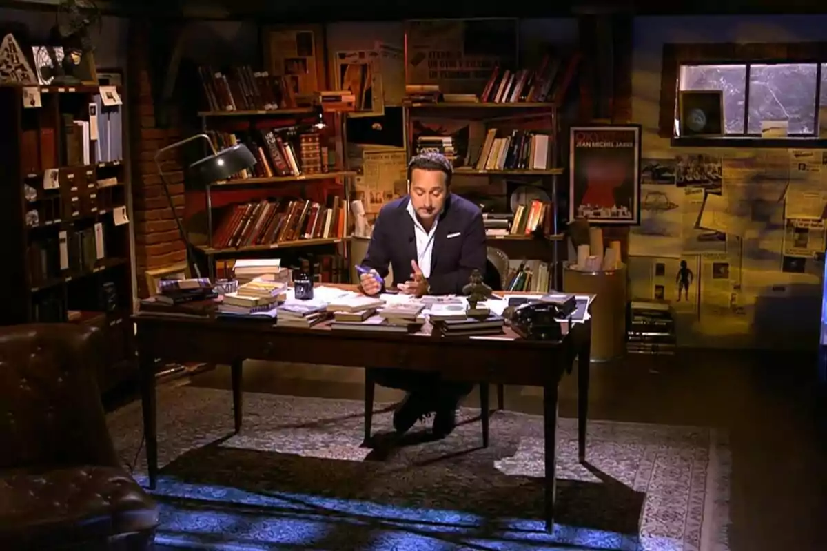 Iker Jiménez sentado en un escritorio antiguo rodeado de libros y papeles en una biblioteca con luz cálida y ambiente acogedor en Cuarto Milenio