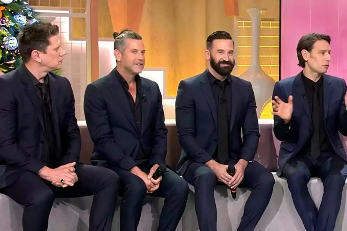 Cuatro hombres del grupo Il Divo vestidos con trajes oscuros están sentados en un sofá durante una entrevista en un set de televisión decorado con un árbol de Navidad y jarrones en Fiesta.