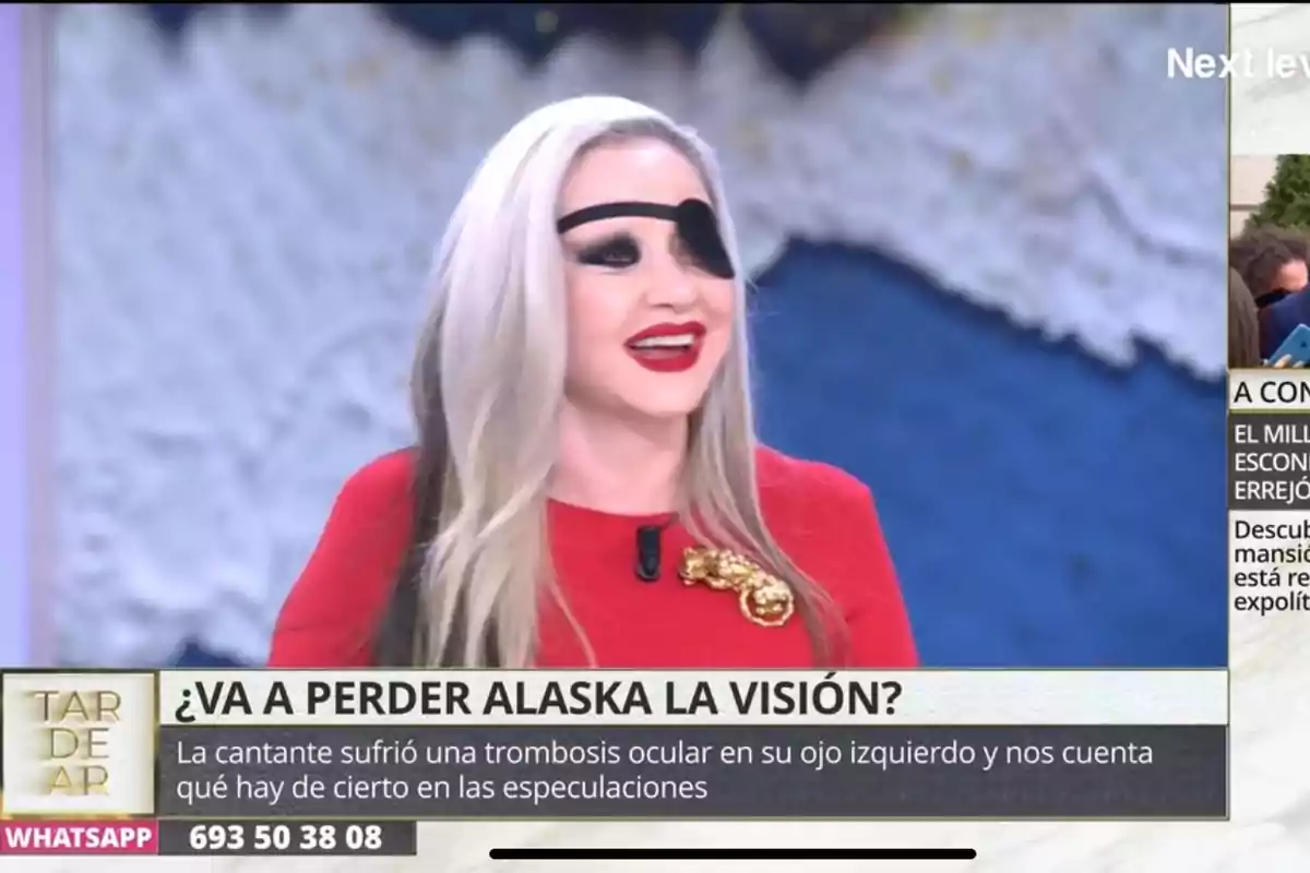 Alaska con un parche en el ojo izquierdo y cabello rubio sonríe en un programa de televisión con un texto en pantalla que menciona una trombosis ocular. Alaska con un parche en el ojo izquierdo y cabello rubio sonríe en un programa de televisión con un texto en pantalla que menciona una trombosis ocular.