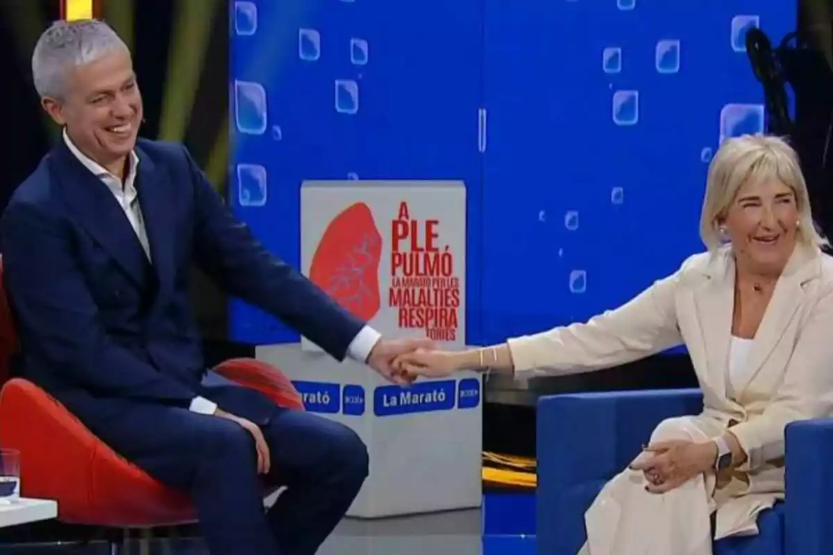 Dues persones somriuen mentre es prenen de la mà en un programa de televisió amb un cartell de fons que diu 