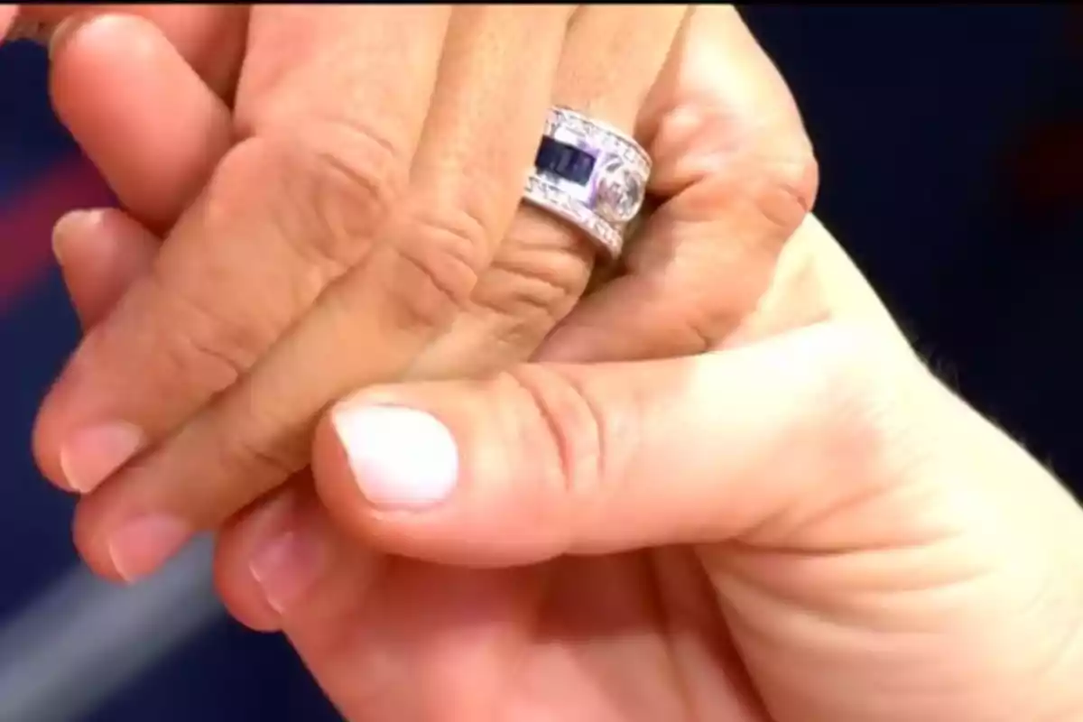 Dos manos entrelazadas mostrando un anillo plateado con detalles oscuros. Dos manos entrelazadas mostrando un anillo plateado con detalles oscuros.