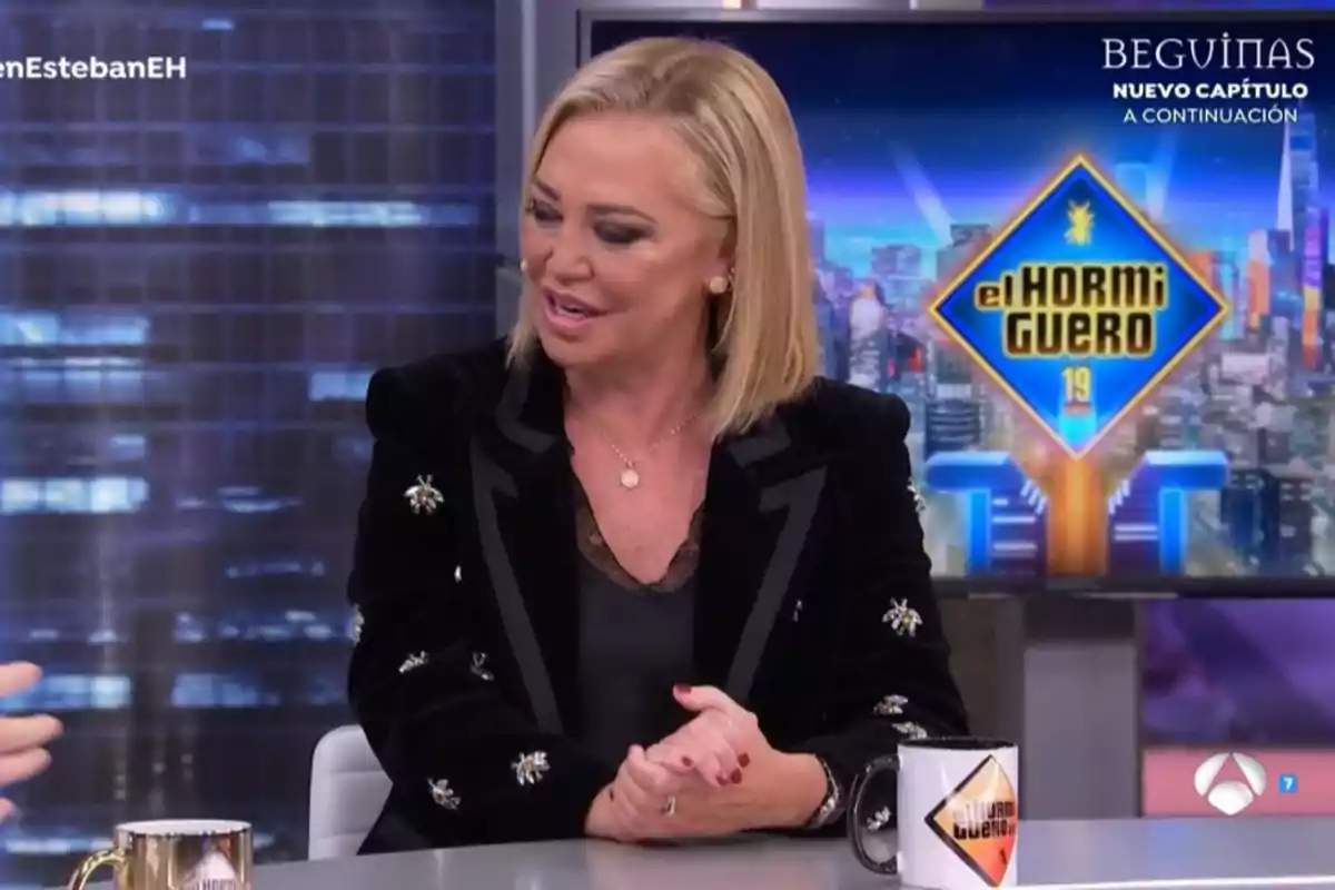 Belén Esteban con chaqueta negra decorada con detalles brillantes está sentada en un plató de televisión con el logo de Belén Esteban con chaqueta negra decorada con detalles brillantes está sentada en un plató de televisión con el logo de
