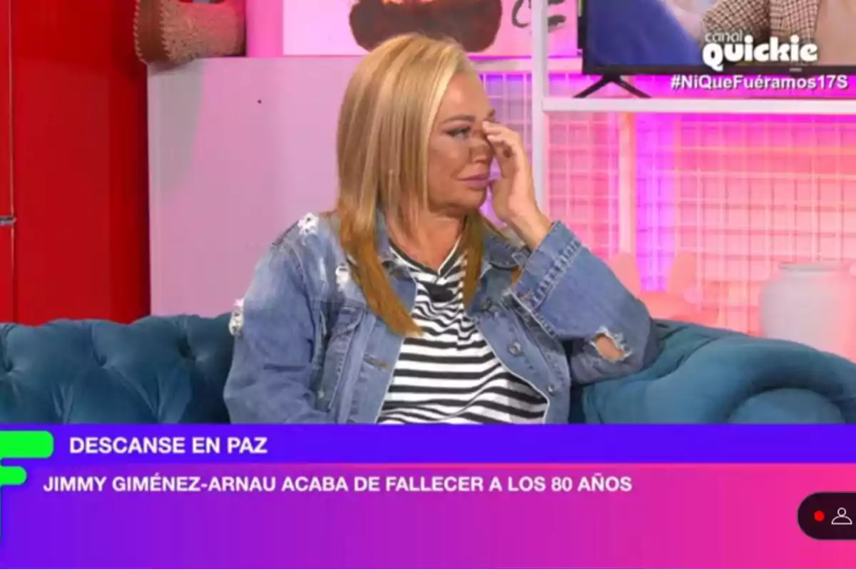 Belén Esteban sentada en un sofá azul, con una chaqueta de mezclilla y camiseta de rayas, parece estar emocionada mientras se toca la cara; en la pantalla se lee Belén Esteban sentada en un sofá azul, con una chaqueta de mezclilla y camiseta de rayas, parece estar emocionada mientras se toca la cara; en la pantalla se lee
