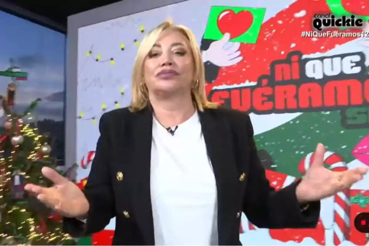 Belén Esteban con cabello rubio y chaqueta negra está frente a un fondo decorado con motivos navideños y un cartel que dice Belén Esteban con cabello rubio y chaqueta negra está frente a un fondo decorado con motivos navideños y un cartel que dice
