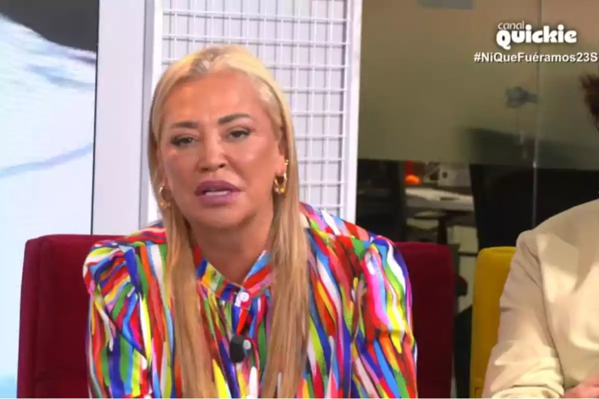 Belén Esteban con cabello rubio y una blusa de colores brillantes está sentada en un estudio de televisión con el logo de Belén Esteban con cabello rubio y una blusa de colores brillantes está sentada en un estudio de televisión con el logo de