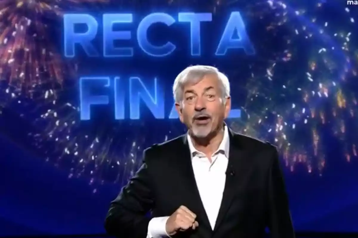Carlos Sobera de cabello canoso y barba está hablando frente a un fondo con fuegos artificiales y las palabras Carlos Sobera de cabello canoso y barba está hablando frente a un fondo con fuegos artificiales y las palabras