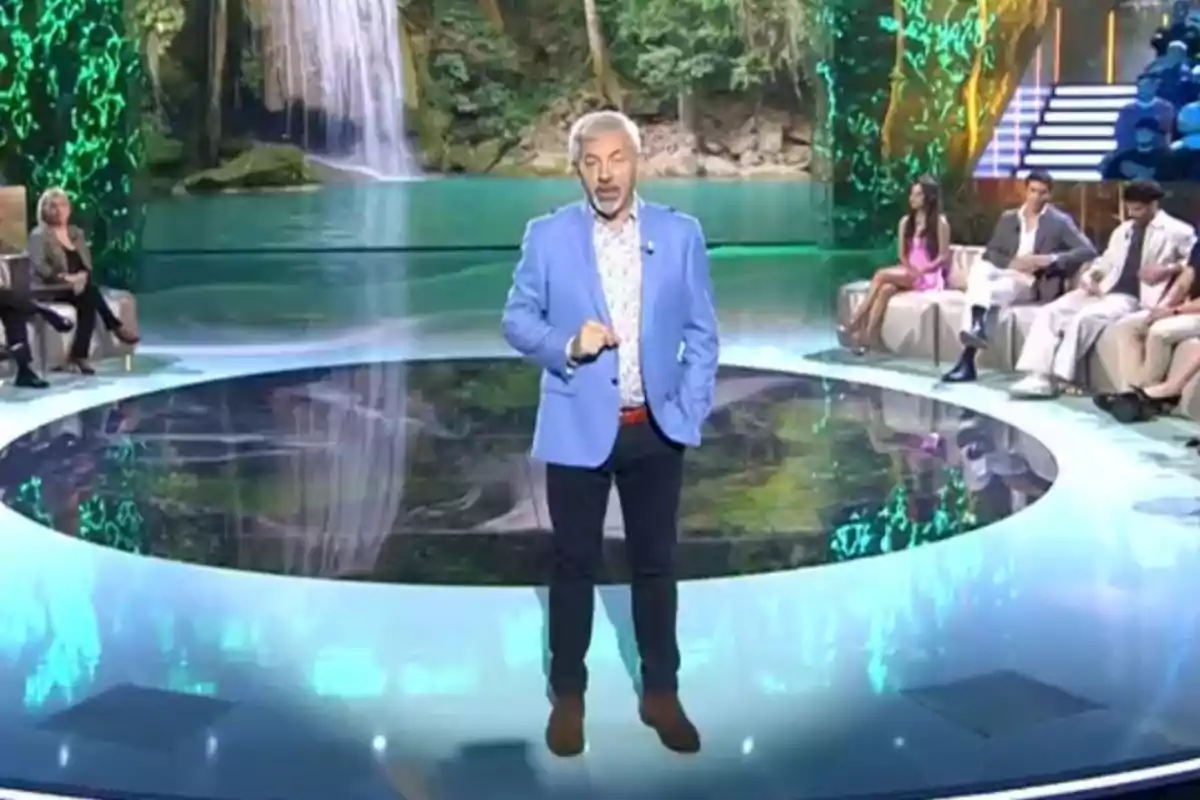 Carlos Sobera está de pie en un escenario circular con un fondo de cascada y vegetación, rodeado de varias personas sentadas en un plató iluminado. Carlos Sobera está de pie en un escenario circular con un fondo de cascada y vegetación, rodeado de varias personas sentadas en un plató iluminado.