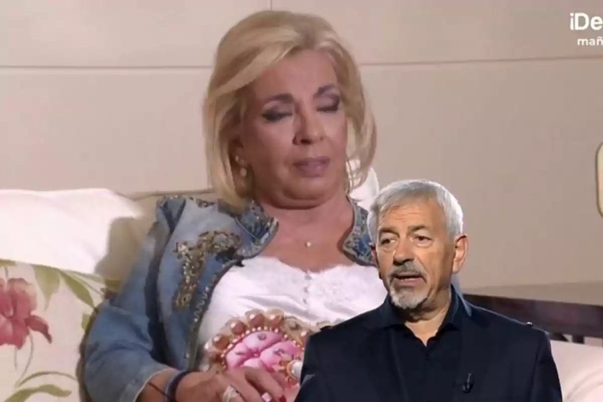 Carmen Borrego con chaqueta decorada está sentada en un sofá con los ojos cerrados mientras Carlos Sobera mayor con barba y cabello canoso aparece en primer plano.