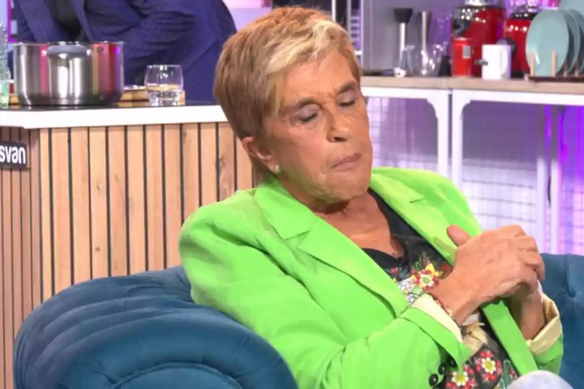 Una persona amb jaqueta verda està asseguda en un sofà blau en un ambient de cuina. Una persona amb jaqueta verda està asseguda en un sofà blau en un ambient de cuina.