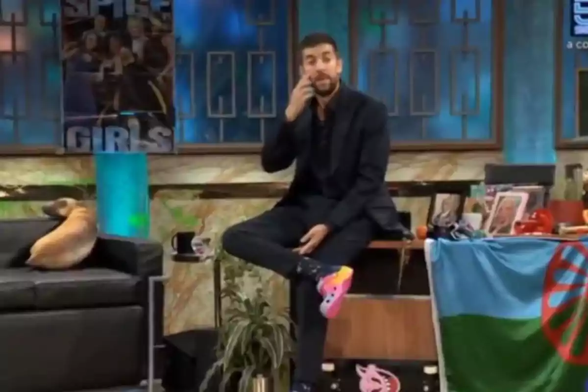 David Broncano sentado en un set de televisión con un póster de las Spice Girls al fondo, un perro en el sofá y una bandera sobre la mesa. David Broncano sentado en un set de televisión con un póster de las Spice Girls al fondo, un perro en el sofá y una bandera sobre la mesa.