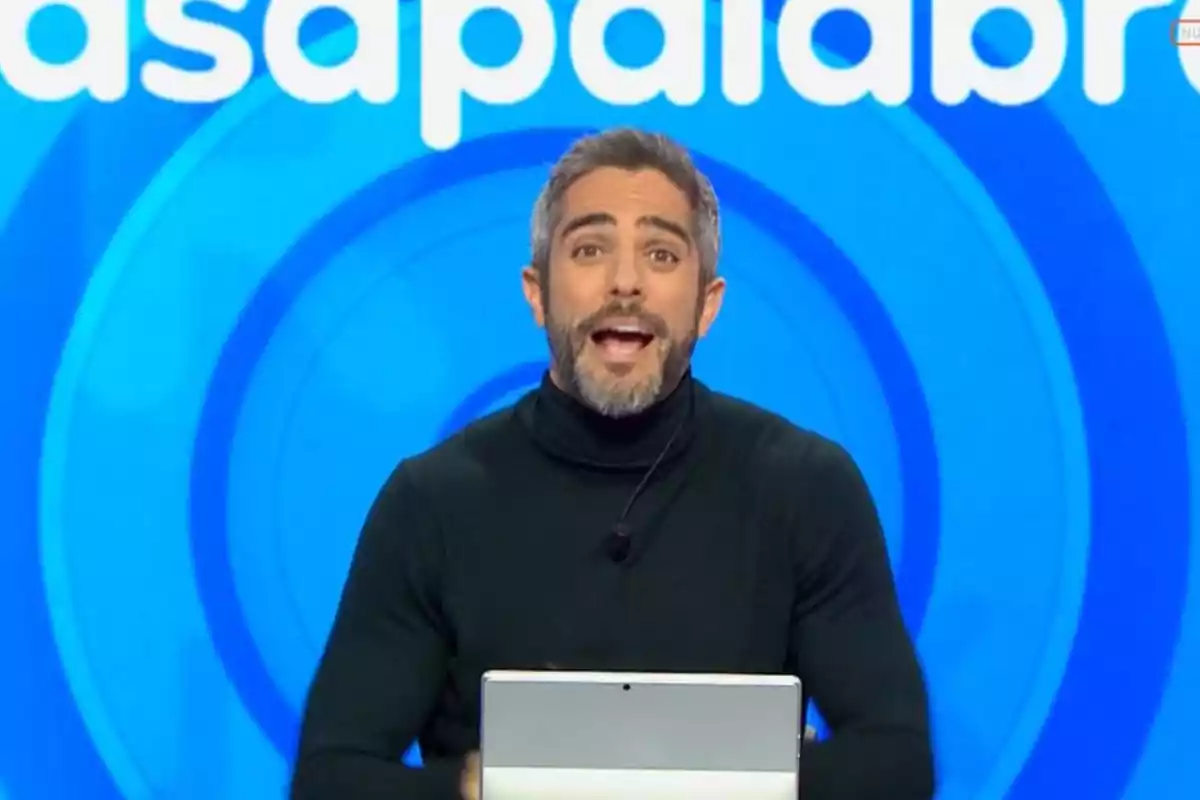 Un presentador de televisió amb barba i cabell canós està parlant davant d'una pantalla blava amb lletres blanques. Un presentador de televisió amb barba i cabell canós està parlant davant d'una pantalla blava amb lletres blanques.