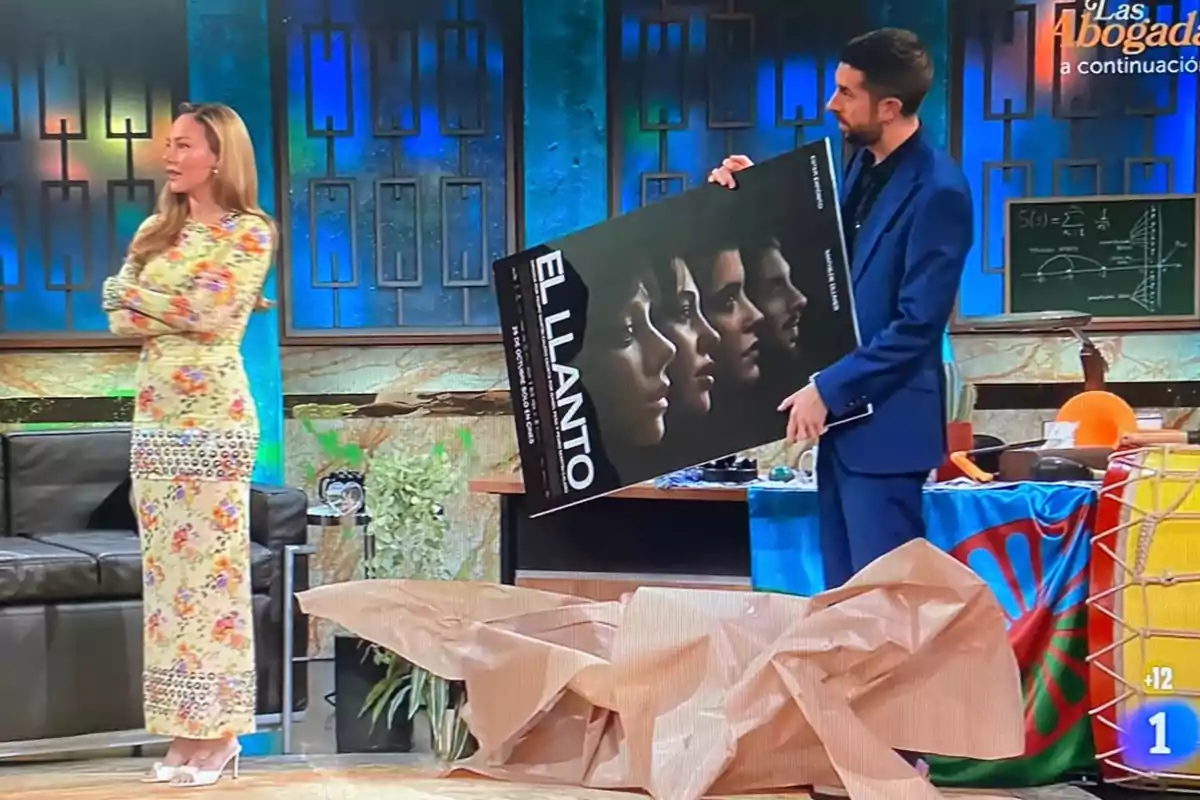 Ester Expósito con un vestido floral está de pie con los brazos cruzados mientras David Broncano sostiene un cartel que dice Ester Expósito con un vestido floral está de pie con los brazos cruzados mientras David Broncano sostiene un cartel que dice