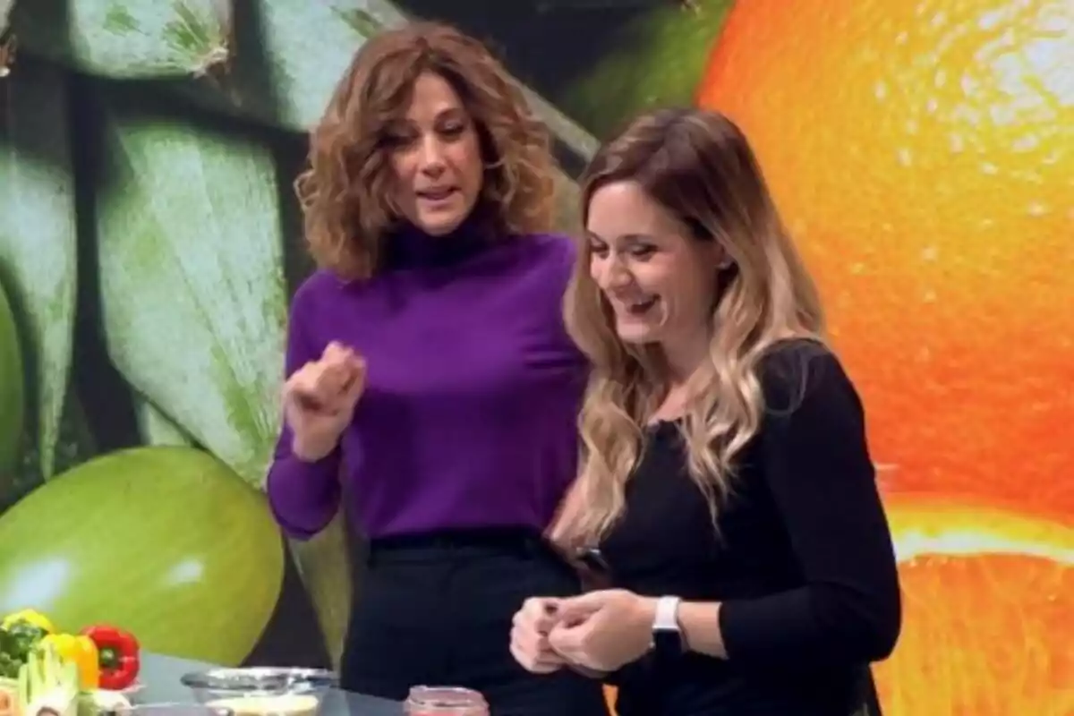 Dos mujeres sonríen mientras cocinan juntas en un programa de cocina con un fondo de frutas y verduras. Dos mujeres sonríen mientras cocinan juntas en un programa de cocina con un fondo de frutas y verduras.