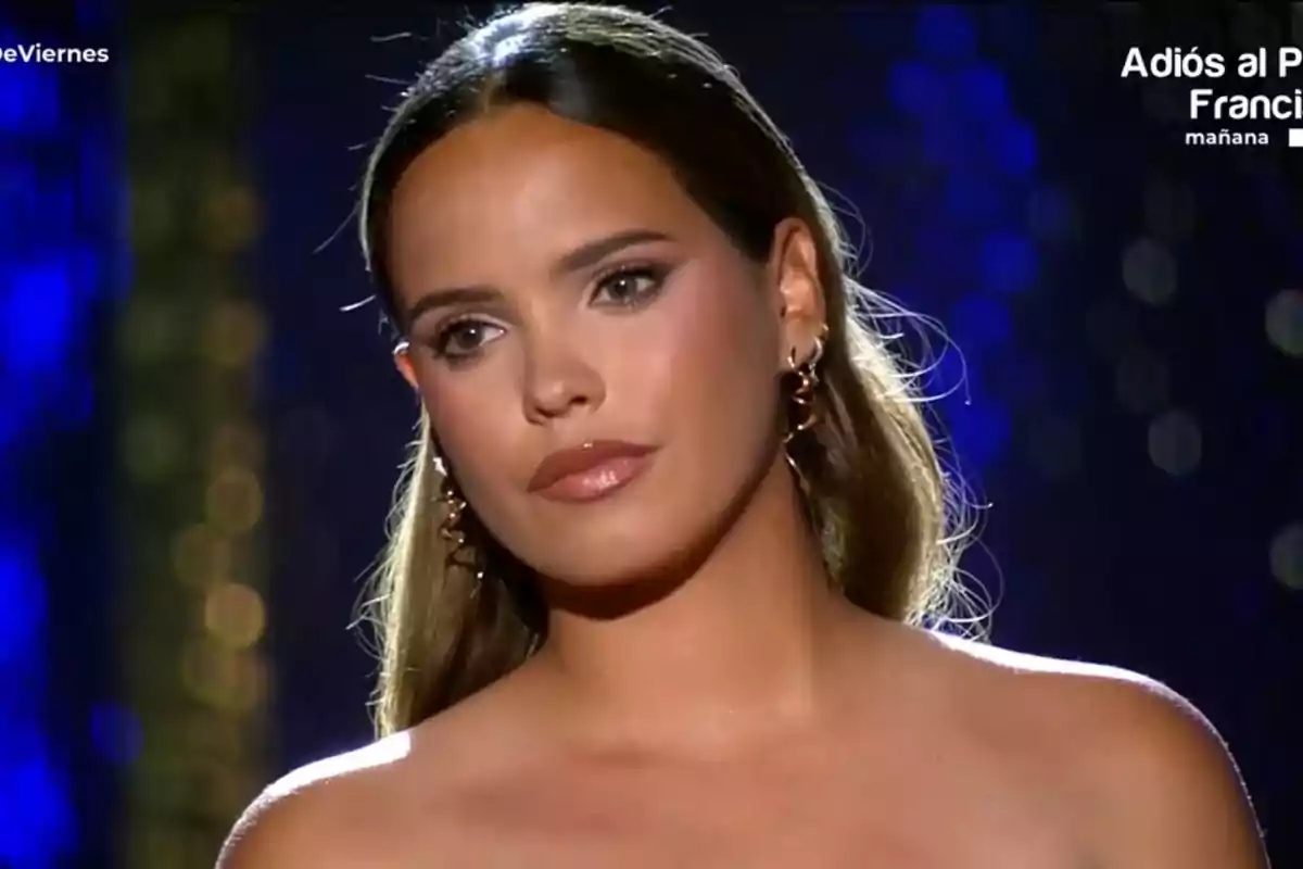 Gloria Camila con cabello largo y oscuro, usando aretes dorados, aparece en un escenario iluminado con luces de fondo desenfocadas. Gloria Camila con cabello largo y oscuro, usando aretes dorados, aparece en un escenario iluminado con luces de fondo desenfocadas.