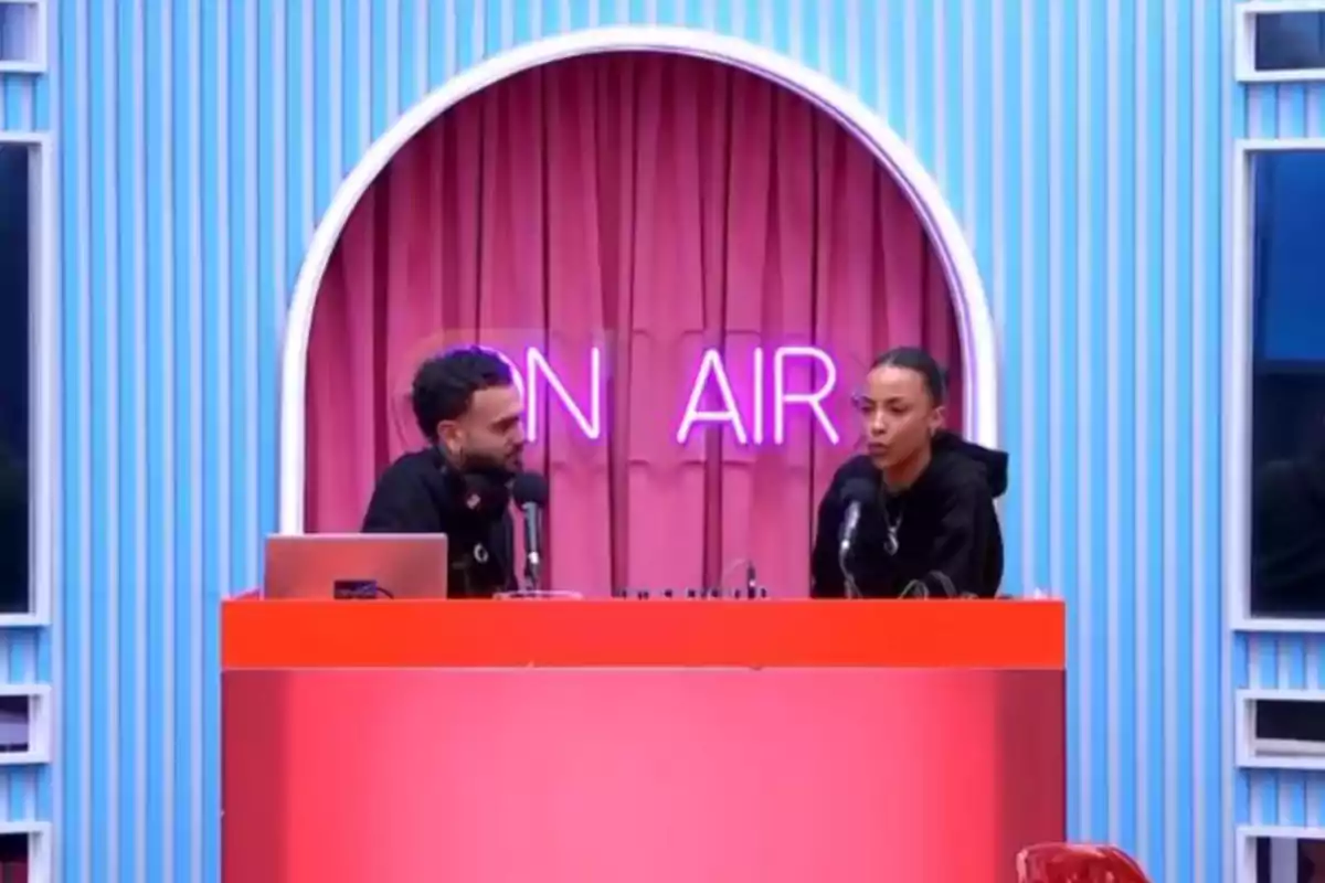 Dues persones conversen en un estudi d'enregistrament amb un rètol de “ON AIR” il·luminat al fons.
