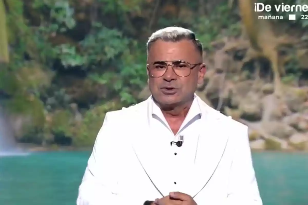 Jorge Javier Vázquez con gafas y cabello canoso está vestido con un traje blanco, de pie frente a un fondo natural con vegetación y agua. Jorge Javier Vázquez con gafas y cabello canoso está vestido con un traje blanco, de pie frente a un fondo natural con vegetación y agua.