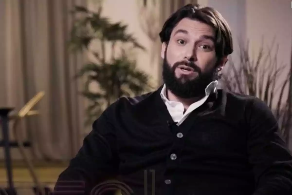 Jota Peleteiro con barba y cabello oscuro está sentado en una silla, vistiendo un suéter negro y camisa blanca, con plantas y cortinas de fondo. Jota Peleteiro con barba y cabello oscuro está sentado en una silla, vistiendo un suéter negro y camisa blanca, con plantas y cortinas de fondo.