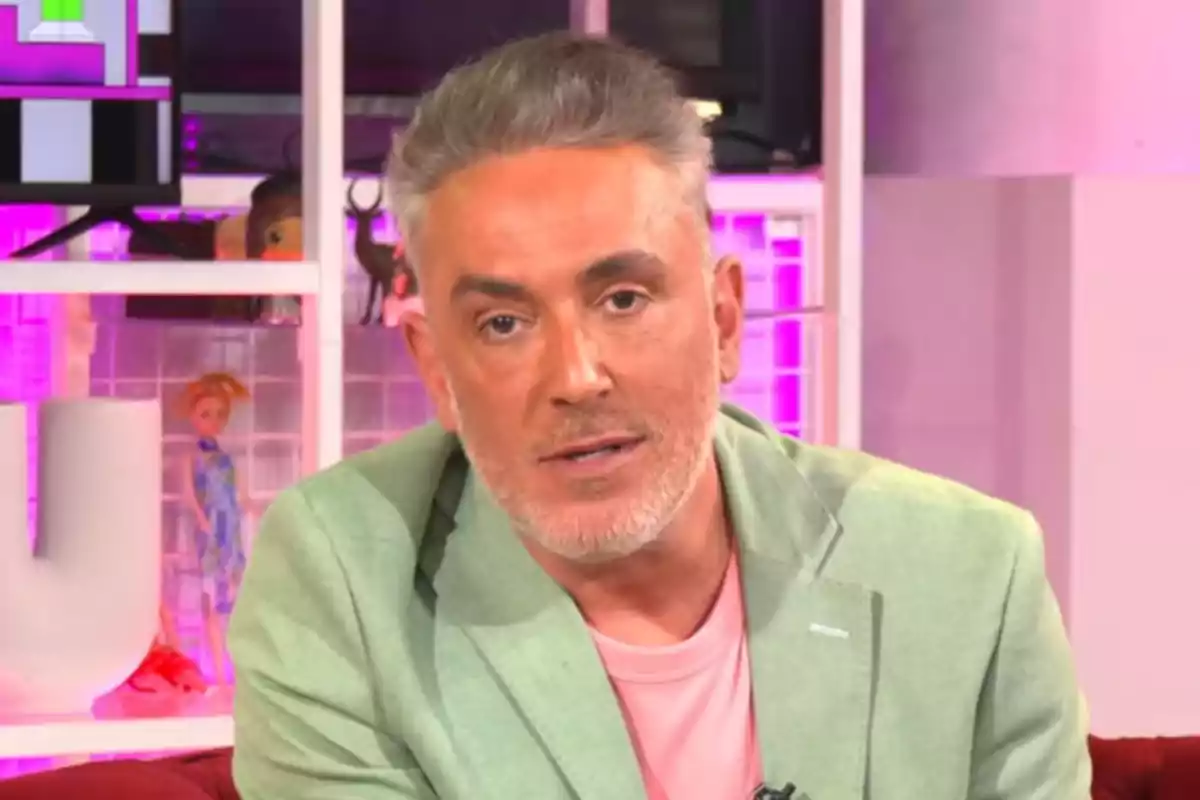 Kiko Hernández con cabello canoso y barba corta lleva una chaqueta verde y camiseta rosa, sentado en un entorno con iluminación rosa y objetos decorativos en el fondo.