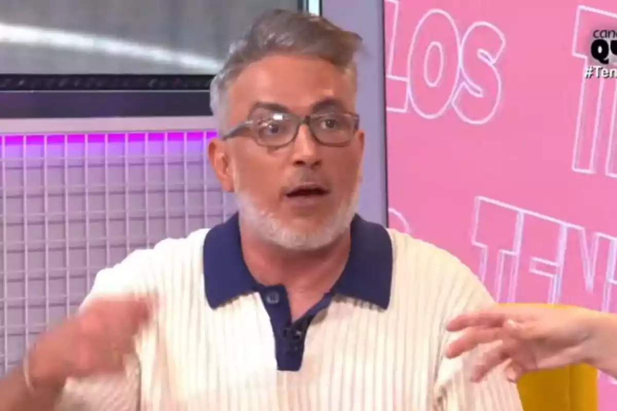 Kiko Hernández con gafas y barba canosa está hablando en un programa de televisión con fondo rosa y letras blancas.