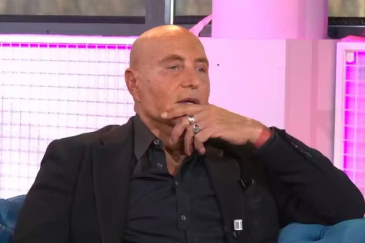 Un home calb amb vestit fosc està assegut en un sofà blau amb una expressió pensativa i la mà a la barbeta, en un entorn amb llums roses.