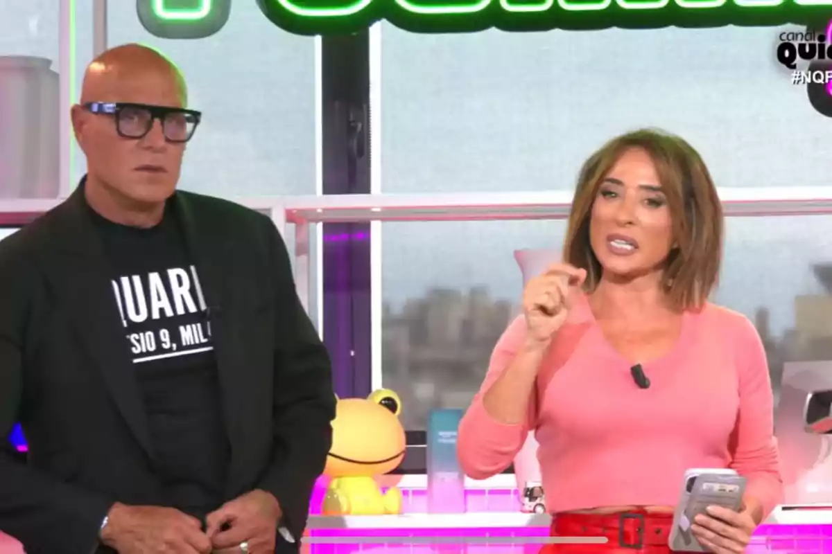Kiko Matamoros y María Patiño están de pie en un set de televisión, Kiko Matamoros lleva gafas y una camiseta negra, mientras que María Patiño viste una blusa rosa y sostiene un teléfono móvil. Kiko Matamoros y María Patiño están de pie en un set de televisión, Kiko Matamoros lleva gafas y una camiseta negra, mientras que María Patiño viste una blusa rosa y sostiene un teléfono móvil.