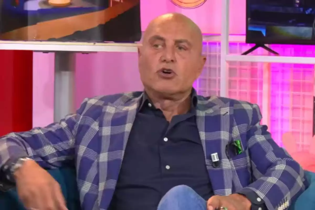 Un home calb amb jaqueta de quadres blaus i camisa fosca està assegut en un sofà en un estudi de televisió amb llums de colors al fons.