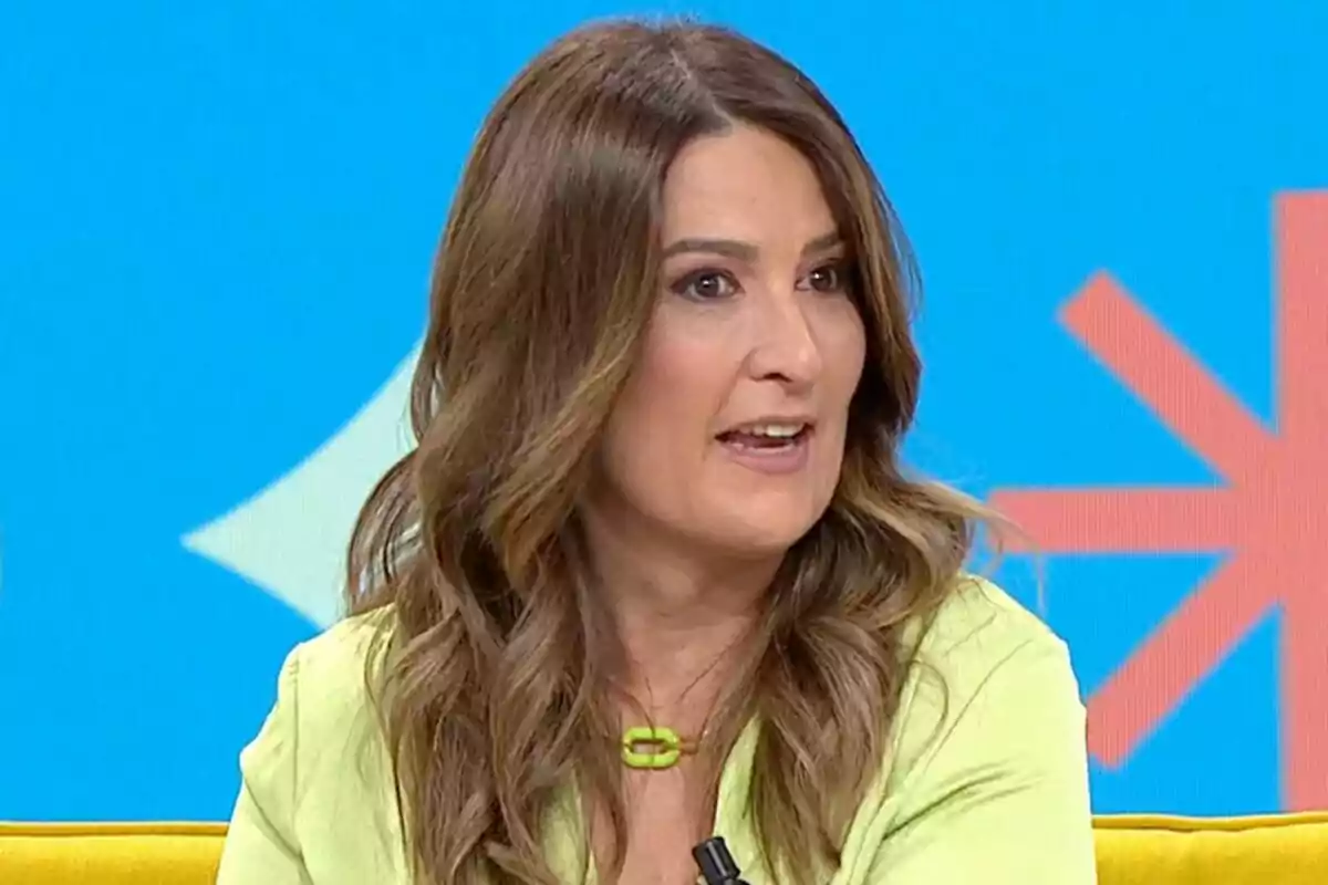 Una mujer con cabello castaño y ondulado hablando en un programa de televisión con un fondo azul y detalles geométricos.