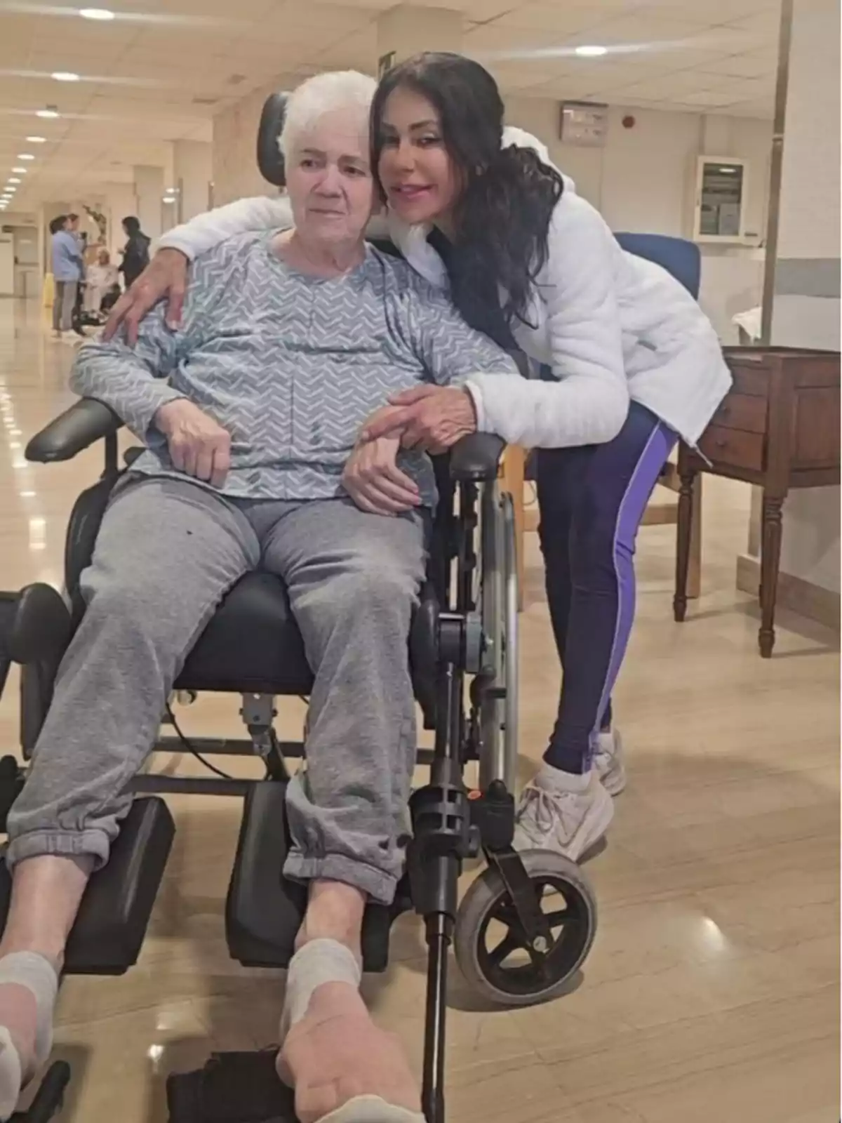 La madre de Maite Galdeano está sentada en una silla de ruedas mientras Maite la abraza y sonríe en un pasillo iluminado. La madre de Maite Galdeano está sentada en una silla de ruedas mientras Maite la abraza y sonríe en un pasillo iluminado.